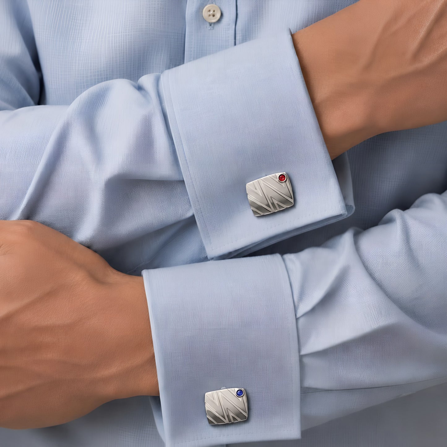 Bandor Essence Cufflinks in solid platinum worn on a light blue dress shirt, arms crossed: luxury men's cufflinks set with a natural ruby on one cuff and a blue sapphire on the other.